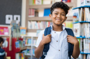 child holding their backpack straps and smiling while standing in a school library