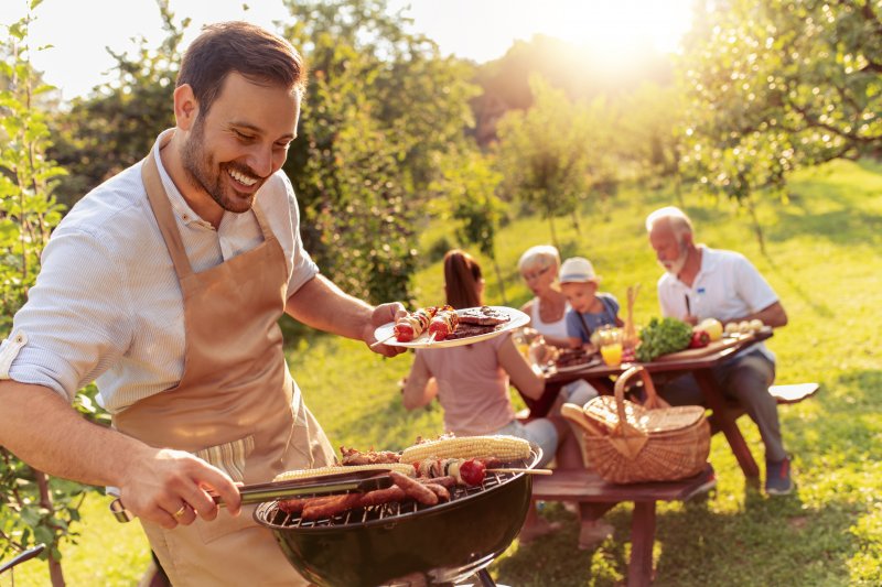 Man smiling while using tongs to pick up a hot dog off a grill with kebabs, hot dogs, and corn on the cob at a barbecue with his family in the background