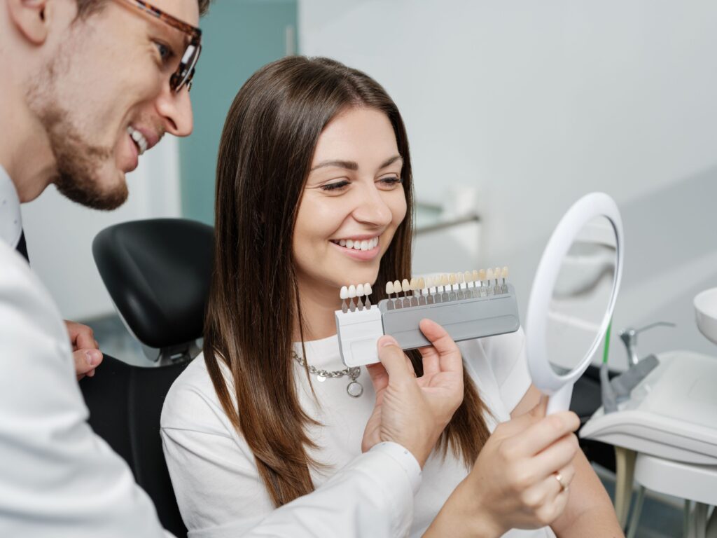 Woman in dental chair smiling at reflection with dentist holding shade guide to her teeth